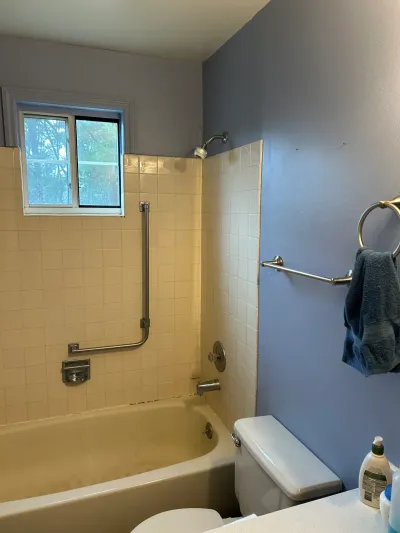 A dated bathroom with a cream tile tub surround, blue walls, and a window above the tub before renovation.