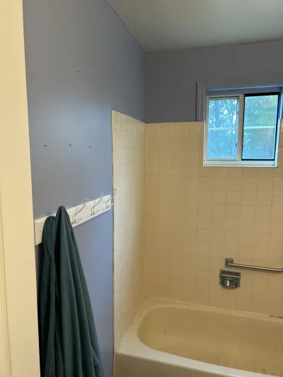Dated bathroom with beige tile tub surround, blue-gray walls, and a window above the tub, before renovation.