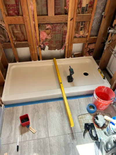 A new shower pan sits amid exposed wall framing and insulation during a bathroom remodel, with tools scattered on the tile fl