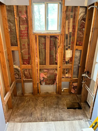Gutted bathroom mid-renovation showing exposed studs, insulation, and plumbing rough-in near a window.