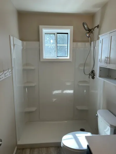 White shower stall with corner shelves and a window, next to a toilet and small wall cabinet.