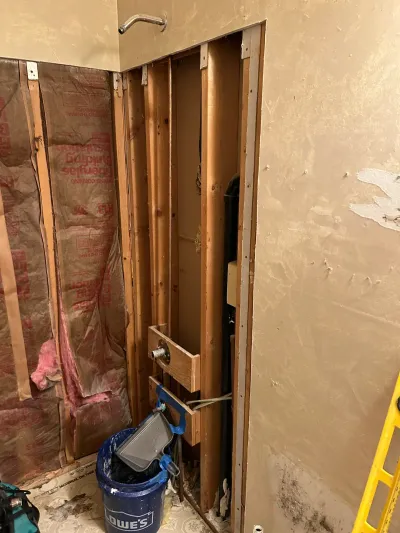 Bathroom wall opened to studs during renovation, showing insulation, framing, and shower plumbing rough-in.