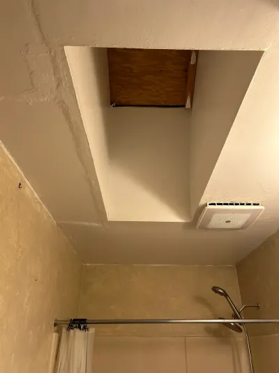 Close-up of a bathroom ceiling with an exposed plywood access panel and an exhaust fan vent above a shower.