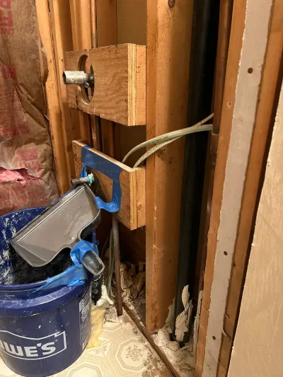 Open bathroom wall showing exposed studs, plumbing rough-in, and a bucket on old tile flooring during renovation.