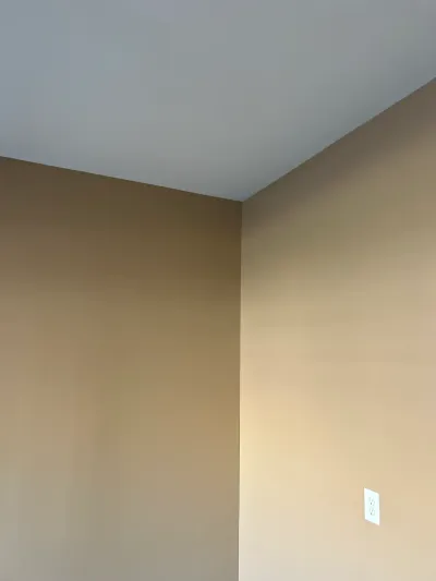 Close-up of a freshly painted tan wall corner meeting a light gray ceiling, with an electrical outlet.