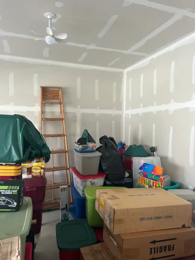 A garage with unfinished taped drywall walls and ceiling, cluttered with storage totes and boxes.