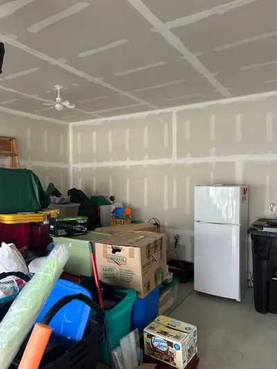 Garage interior with unfinished taped drywall, a white refrigerator, and stacked moving boxes.