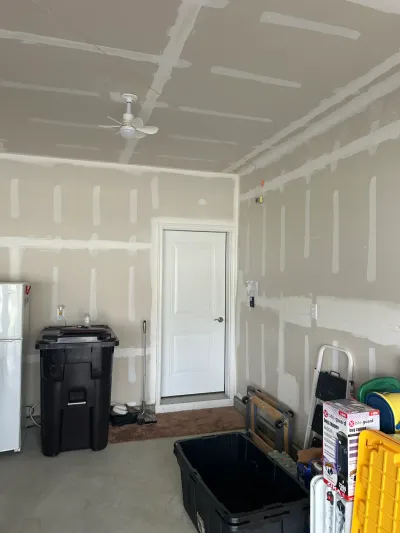 Garage under renovation with taped drywall seams, a ceiling fan, and storage bins on the concrete floor.