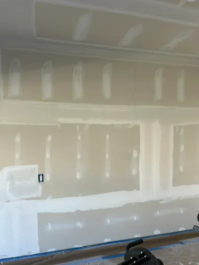 A wall and ceiling mid-drywall finishing, with joint compound patches, tape seams, and a sander on the floor.