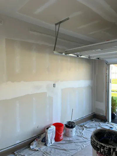 Garage under renovation with taped drywall seams, joint compound buckets, and the door open to the driveway.