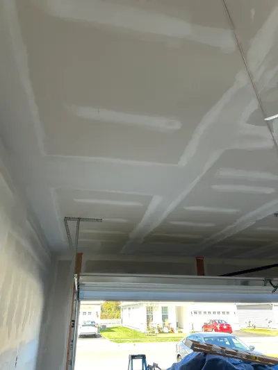 Garage ceiling with taped and mudded drywall seams, mid-installation, open door showing the driveway beyond.