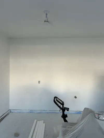 An unfinished room mid-renovation with a partially installed ceiling fan, bare concrete floor, and painting equipment.