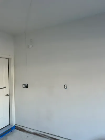 An unfinished room wall with fresh paint, an exposed wire, an outlet, and a white door.