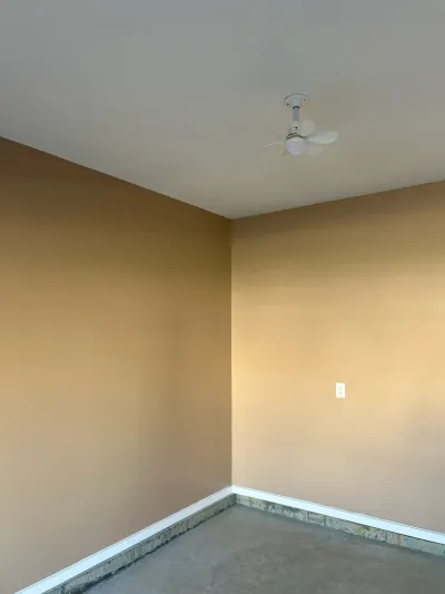 Empty room corner with freshly painted tan walls, white baseboard trim, and a ceiling fan, subfloor exposed.