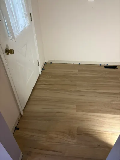 New wood-look vinyl plank flooring being installed in a room, with spacers along the baseboard and a floor vent visible.