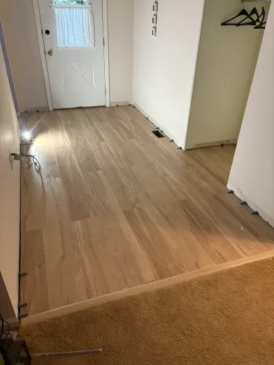 Entryway hallway mid-installation with new oak-look vinyl plank flooring and spacers along the walls, transitioning to carpet