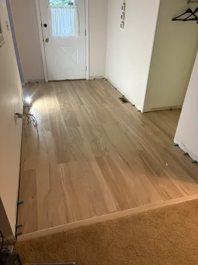 An entryway with newly installed light wood-look vinyl plank flooring, spacers still along the edges, leading to a front door