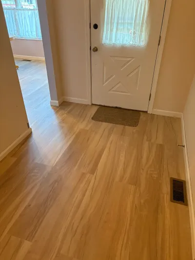 A bright entryway with light wood-look vinyl flooring, a white paneled front door, and a small entry mat.