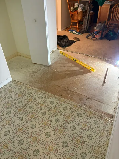 Old vinyl tile flooring partially removed to reveal plywood subfloor, with tools staged for renovation work.