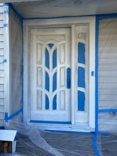 A front door mid-repainting, covered in primer with blue tape protecting the glass panels and siding.
