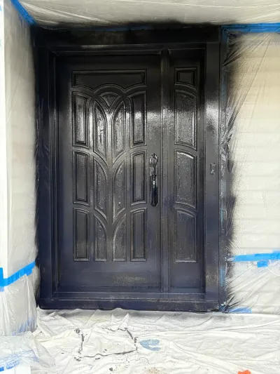 A double entry door mid-spray-painting in matte black, with surrounding walls masked in plastic sheeting.