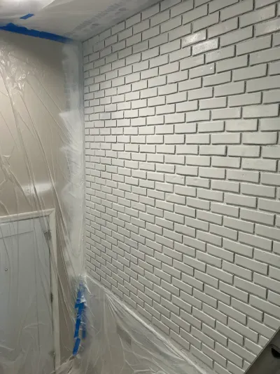 A brick wall freshly painted white, with plastic sheeting and blue tape protecting nearby surfaces during painting.