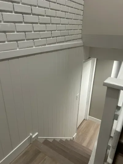 A staircase with white brick-look accent wall and wood paneling leads down to a hallway with plank flooring.