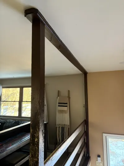 A worn wood beam and post frame an open railing overlooking a lower room, with a window and decor nearby.