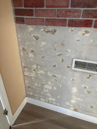 Close-up of a painted brick wall with peeling paint, a wall vent, and new plank flooring near a doorway.