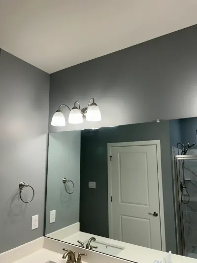 Bathroom vanity with gray walls, a large mirror, and a brushed nickel light fixture reflecting a corner glass shower.