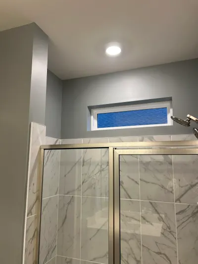 A tiled shower stall with a gold-framed glass door and a small window above it in a gray-painted bathroom.
