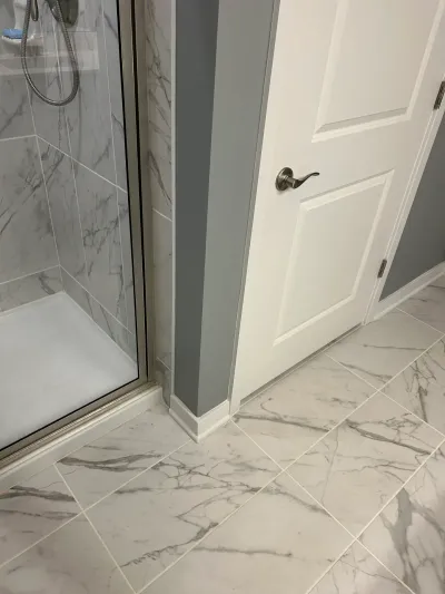 Close-up of a marble-look tiled bathroom corner with a glass shower door and white paneled door.