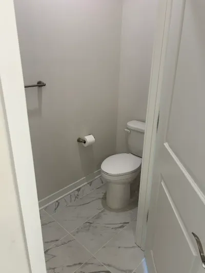 A small finished bathroom with a toilet, diagonal marble-look tile floor, and brushed nickel fixtures.