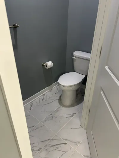 A small bathroom with gray walls, marble-look tile floor, and a toilet, viewed through a doorway.
