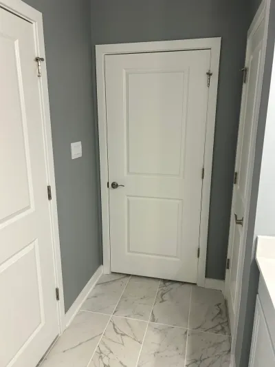A bathroom hallway with gray walls, white paneled doors, and diagonally laid marble-look tile flooring.