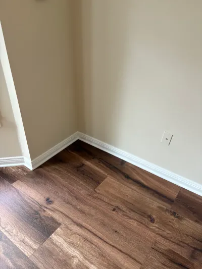 Close-up of a finished room corner with new wood-look flooring, white baseboard, and tan walls.