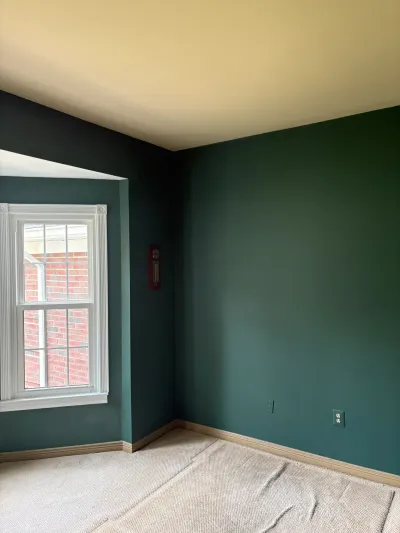 A bedroom with dark green walls, an unfinished cream ceiling, and a window overlooking a brick exterior.