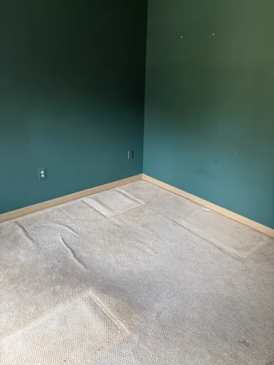 Empty room corner with worn beige carpet and teal walls before renovation.