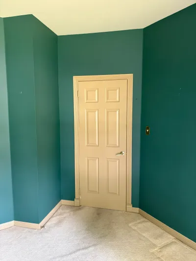 An empty room with teal walls, a beige paneled door, and worn carpet flooring before renovation.