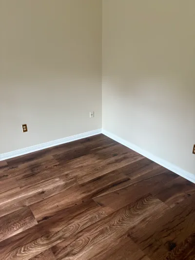 Empty room corner with new dark wood-look vinyl plank flooring and white baseboards against tan walls.
