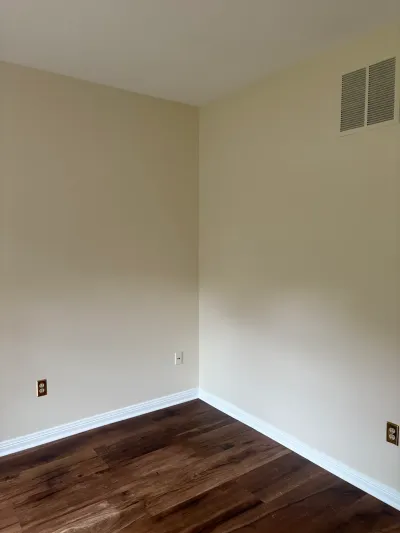 Empty room corner with dark wood flooring, cream walls, white trim, and a ceiling air vent.