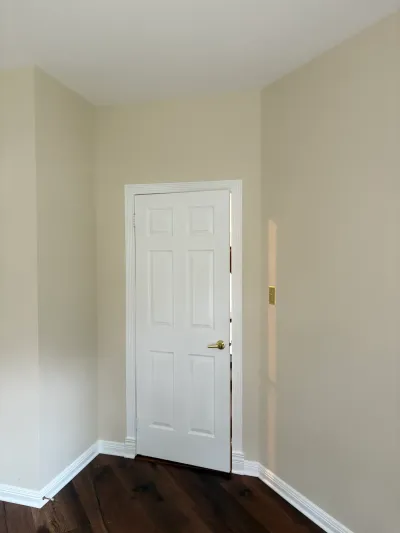 A white paneled door with brass hardware stands ajar in a beige hallway with dark hardwood floors.