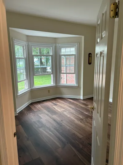 Empty room with dark wood flooring and a bay window overlooking a lawn, seen through an open doorway.