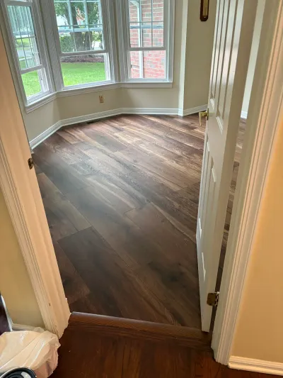 Newly installed dark wood-look plank flooring in a sunlit room with a bay window, seen through an open doorway.