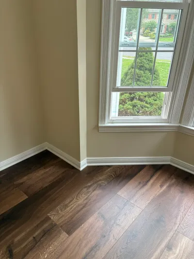 Empty room corner with dark wood flooring and a bay window overlooking a tree-lined street.