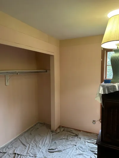 A bedroom mid-painting with peach walls, drop cloths on the floor, and an open closet shelf.