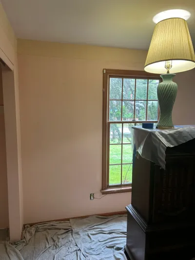 Bedroom with pink walls and a wood-trimmed window, floor and furniture covered in drop cloths before renovation.