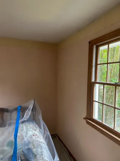 Bedroom corner before painting, with furniture wrapped in plastic near a wood-framed window.