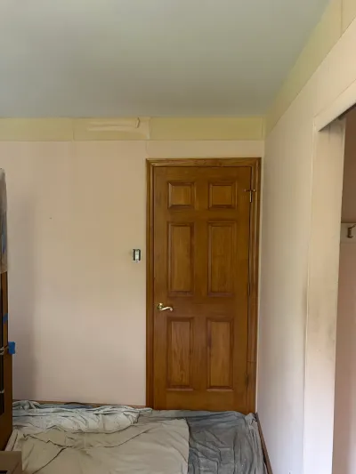 A bedroom mid-renovation with a paneled wood door, taped ceiling edges, and furniture covered by drop cloths.