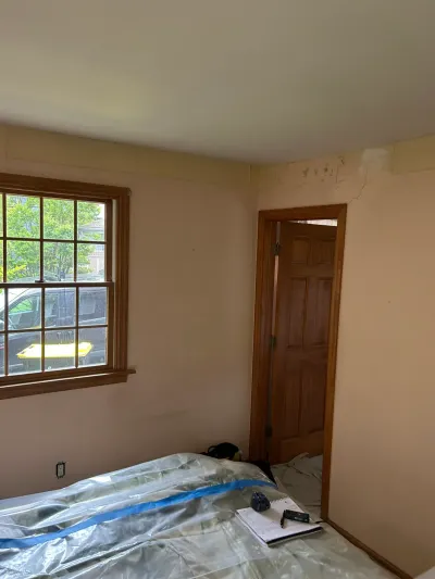 A bedroom with drop-cloth covered furniture, a window, and a doorway showing ceiling cracks before renovation.